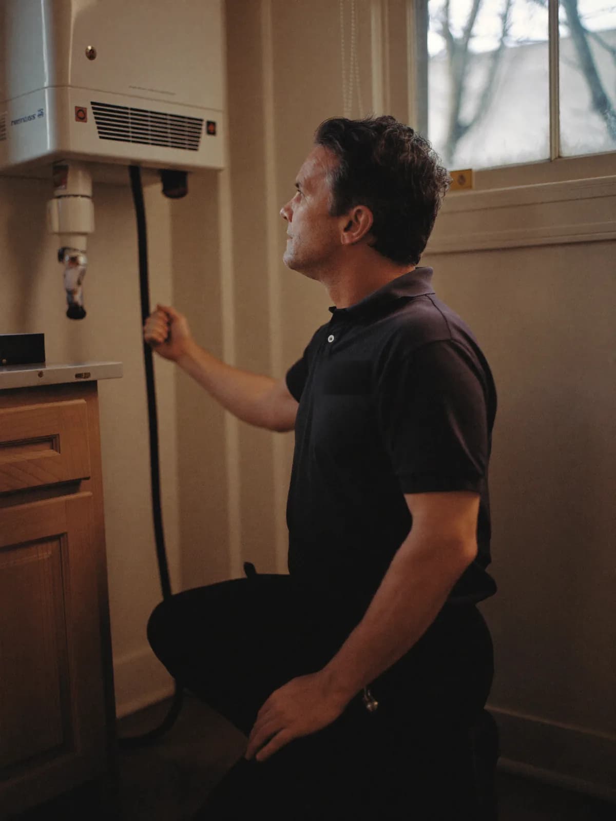 A South East Plumbing engineer kneeling beside a wall-mounted boiler in a domestic kitchen, checking the pipework in warm evening light.